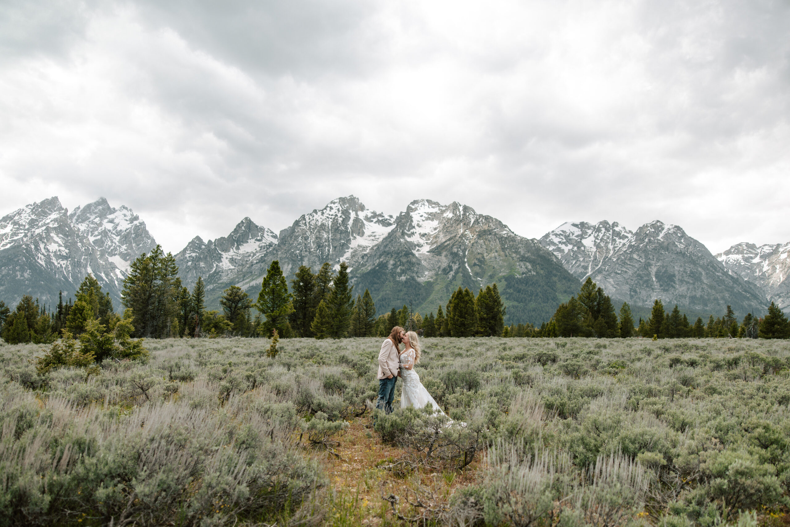 Two People standing in a field in wedding attire with mountains in the background